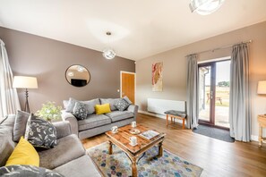 Un salon moderne avec un canapé gris, une table basse et une vue sur l’extérieur, accessible par une porte vitrée.