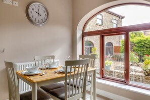 Un coin repas avec une table dressée pour quatre personnes, une horloge murale et une vue sur un bâtiment en pierre, visible à travers une fenêtre.