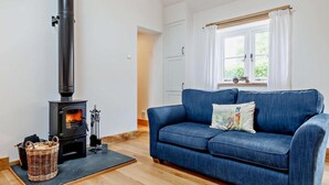 A cozy living room with a blue sofa, a black wood-burning stove, a wicker basket, and a window with curtains. - 1 Bed in Taunton (oc-80693) (North Curry)