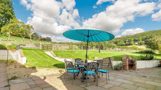 Una veranda esterna con tavolo e sedie, un ombrello blu e un barbecue in mattoni.