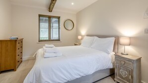 Ein Schlafzimmer mit einem Bett, Nachttischlampen, einer Holzkommode, einem Fenster mit Jalousien und einer Decke mit sichtbaren Holzbalken.