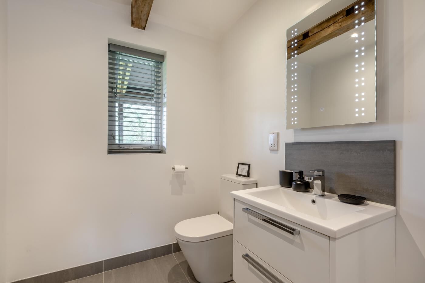 Une salle de bain équipée de toilettes, d’un lavabo, d’un miroir et d’une fenêtre avec des stores.