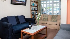 Ein Wohnzimmer mit einem dunkelblauen Sofa, einem beigen Sofa, einem Couchtisch aus Holz mit einer Flasche und Gläsern, einem Bücherregal und einem Fenster mit Vorhängen.