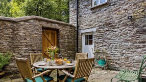 A stone building with a wooden door and window, a round table with chairs, and a small garden area with a watering can. - 4 Bed in Hay-on-wye (oc-bn116) (Hay-on-Wye)