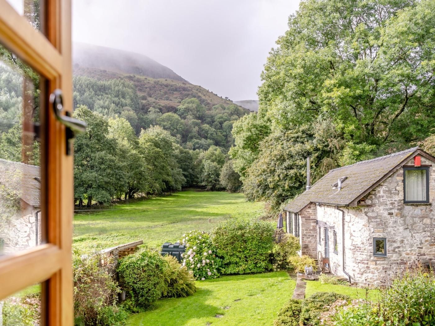 A stone cottage with a garden, surrounded by lush greenery and hills.