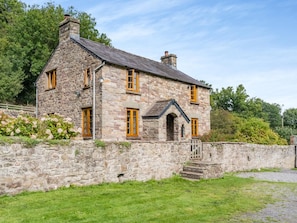 Une maison en pierre avec un toit à deux versants, un petit porche et un mur de pierre qui entoure la propriété.