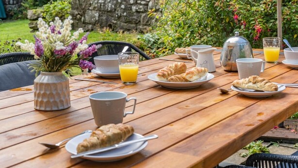 Ein Holztisch, reichhaltig mit Frühstücksleckereien wie Croissants, einer Kanne, Tassen und einem Glas Orangensaft gedeckt.