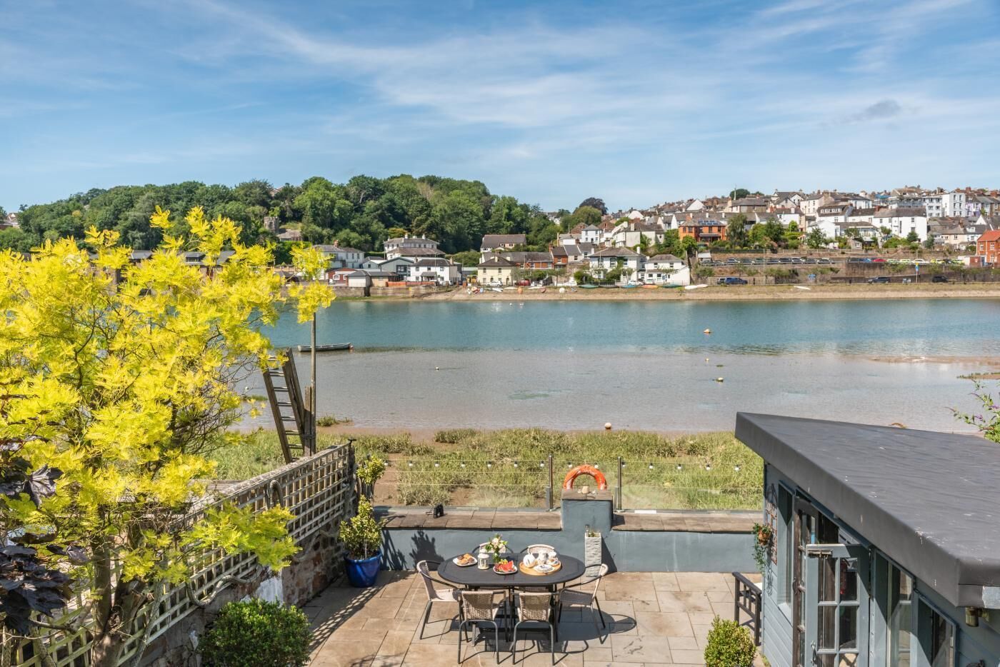 Eine Terrasse mit Tisch und Stühlen mit Blick auf ein Gewässer, ein Baum mit gelben Blättern und der Blick auf eine Stadt am Wasser.