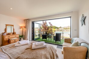 A bedroom with a large bed, a dresser, a mirror, and a view of a balcony with outdoor furniture.