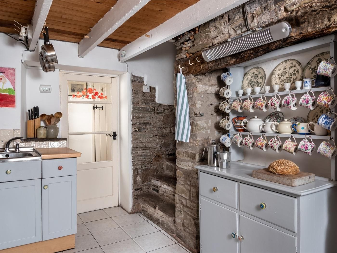 A kitchen with a stone wall, a hanging shelf with teapots, and a door leading to another room.
