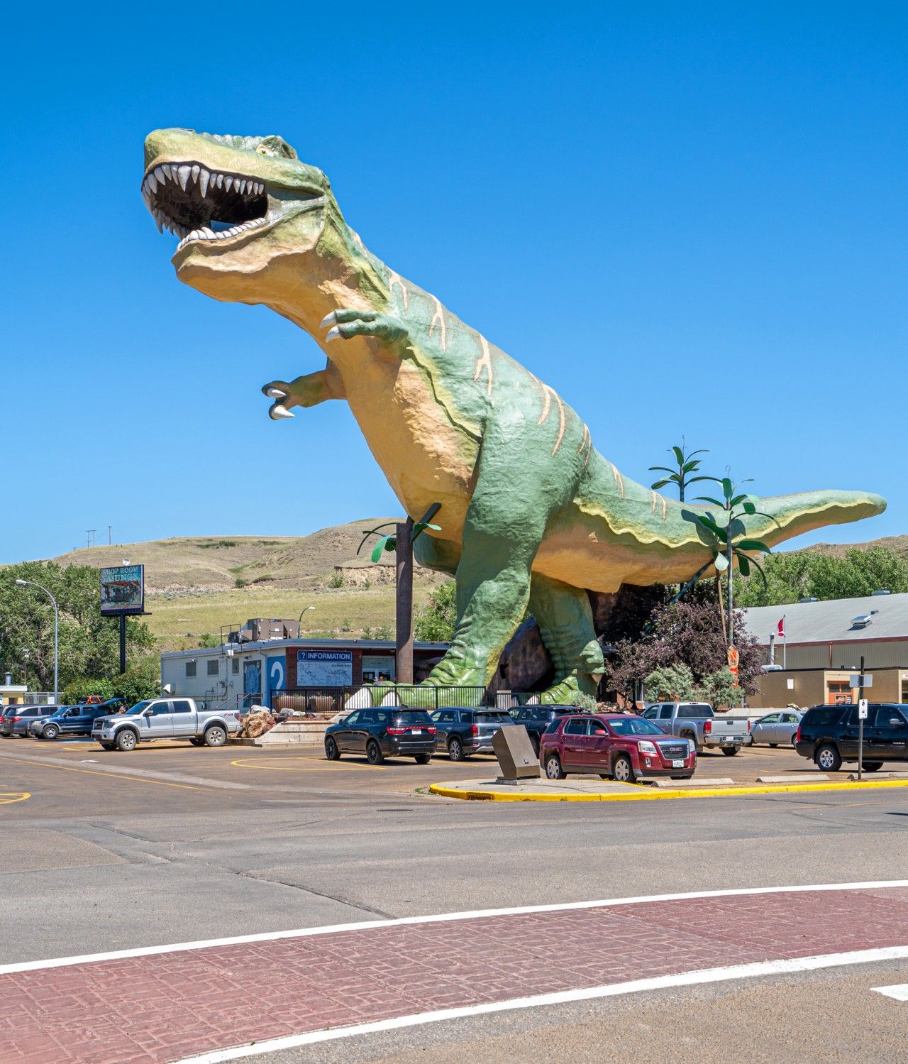 A large dinosaur statue at a roadside intersection with cars and a building in the background.