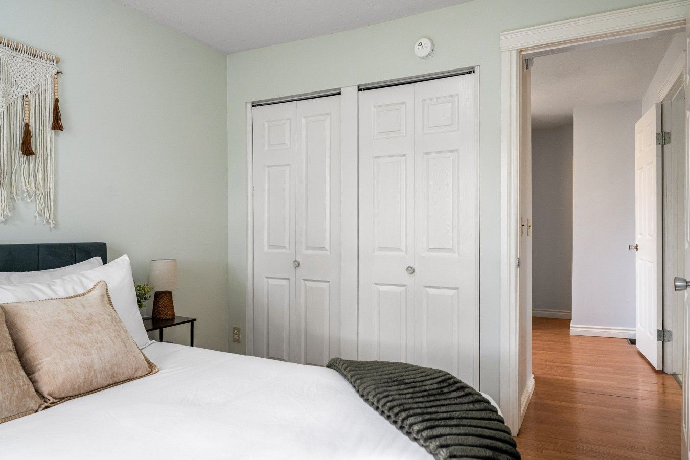 A bedroom with a bed, two white doors, a macrame wall hanging, and a bedside table with a lamp.