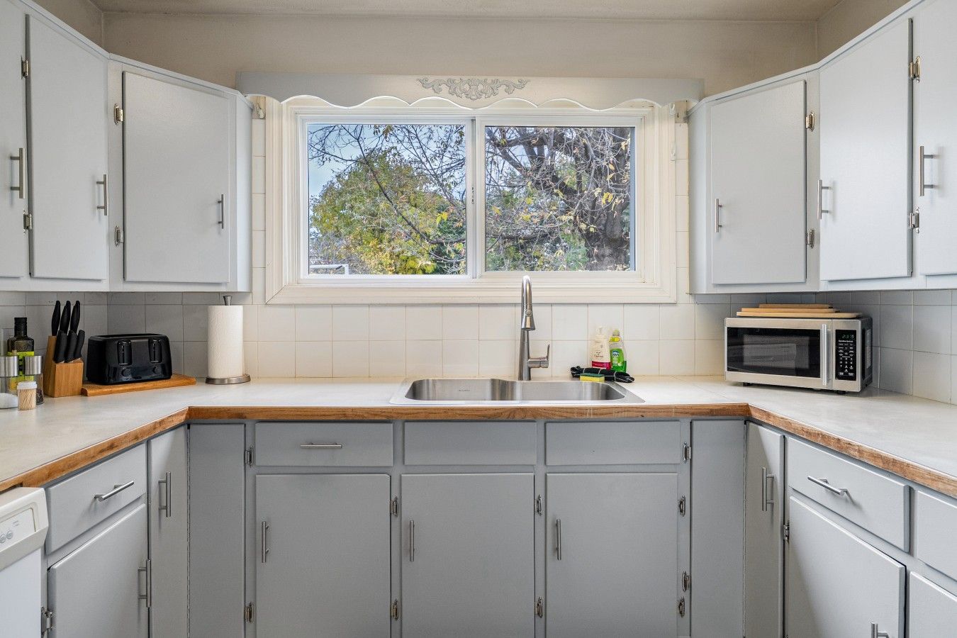 A kitchen with grey cabinets, a stainless steel sink, a microwave, and a window with a view of trees.