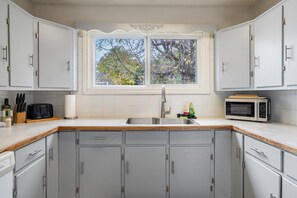 A kitchen with grey cabinets, a stainless steel sink, a microwave, and a window with a view of trees. - The Badlands Basecamp (Drumheller)
