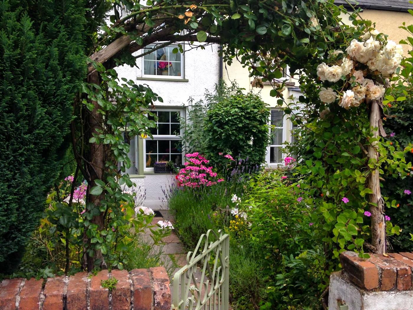 Un jardin avec un mur en briques, une maison blanche et un portail en bois.