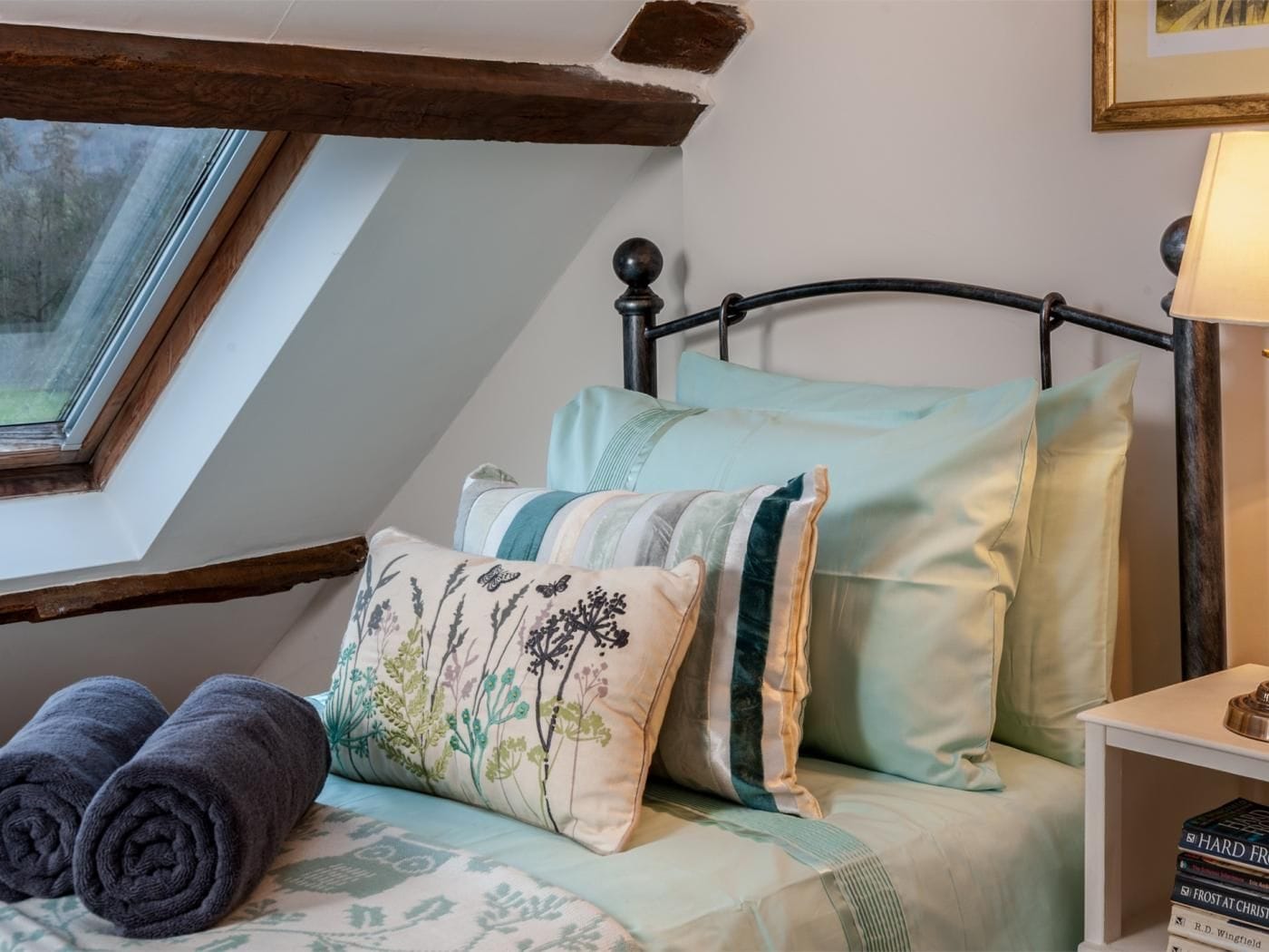 A bedroom with a sloped ceiling, a bed with multiple pillows, a bedside table with books, and a window.