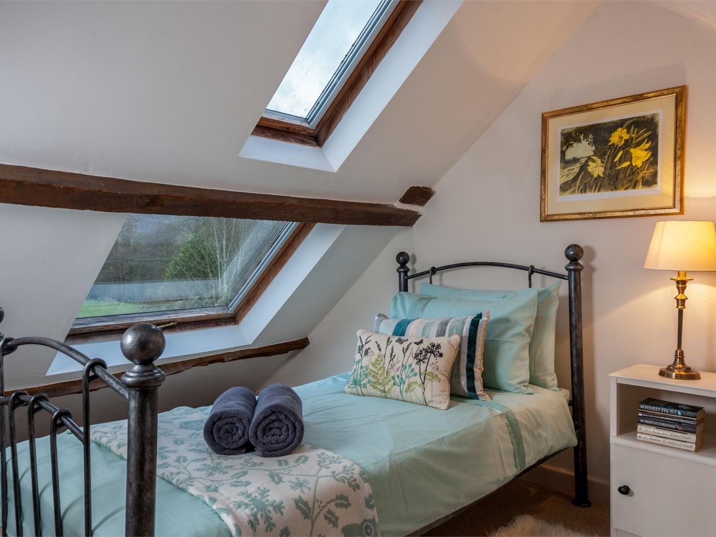 Ein Schlafzimmer mit Dachfenster, einem Bett mit Kissen, einem Nachttisch mit Büchern und einer Lampe.