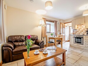 A living room with a brown leather sofa, a wooden coffee table with cups and a vase of flowers, and a kitchen in the background with wooden cabinets and a stove. - 1 Bed in Crickhowell (oc-bn126) (Tretower)