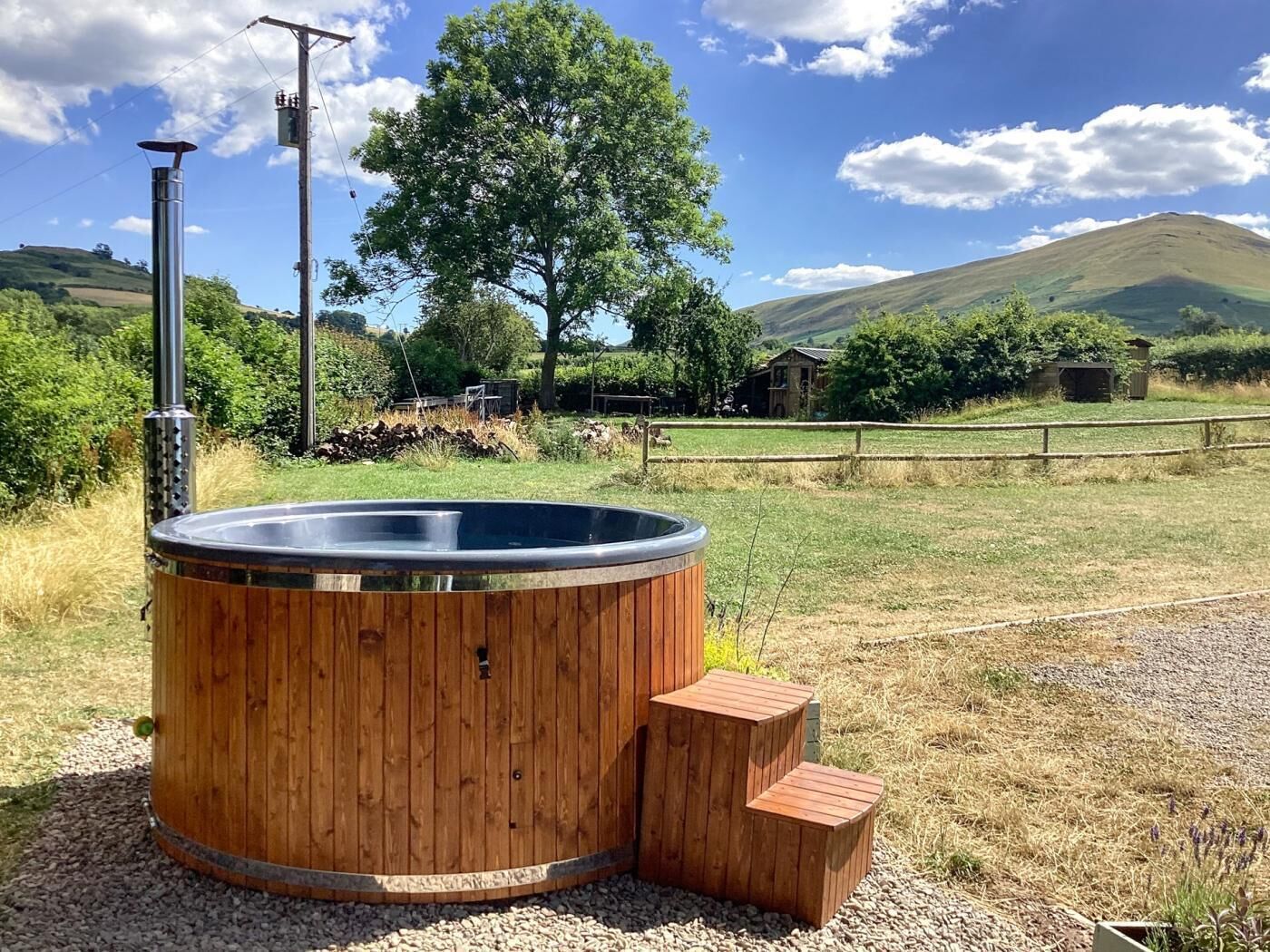 A wooden hot tub with steps, surrounded by a gravel area, with a rural landscape and hills in the background.