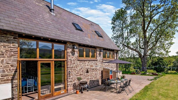 Une maison en pierre avec de grandes fenêtres, une terrasse avec des meubles d’extérieur et un arbre en arrière-plan.