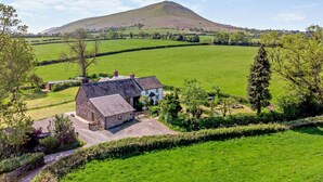 Une maison de campagne aux murs en pierre, entourée de champs verdoyants et avec une colline en arrière-plan.