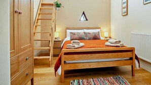 Une chambre avec un lit en bois, une armoire en bois, une échelle en bois, une fenêtre avec des plantes, un radiateur et un tableau encadré au mur.
