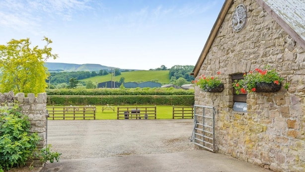A stone building with a gated entrance, flower boxes, and a view of green fields and hills. - 5 Bed in Brecon (oc-bn358) (Llyswen)
