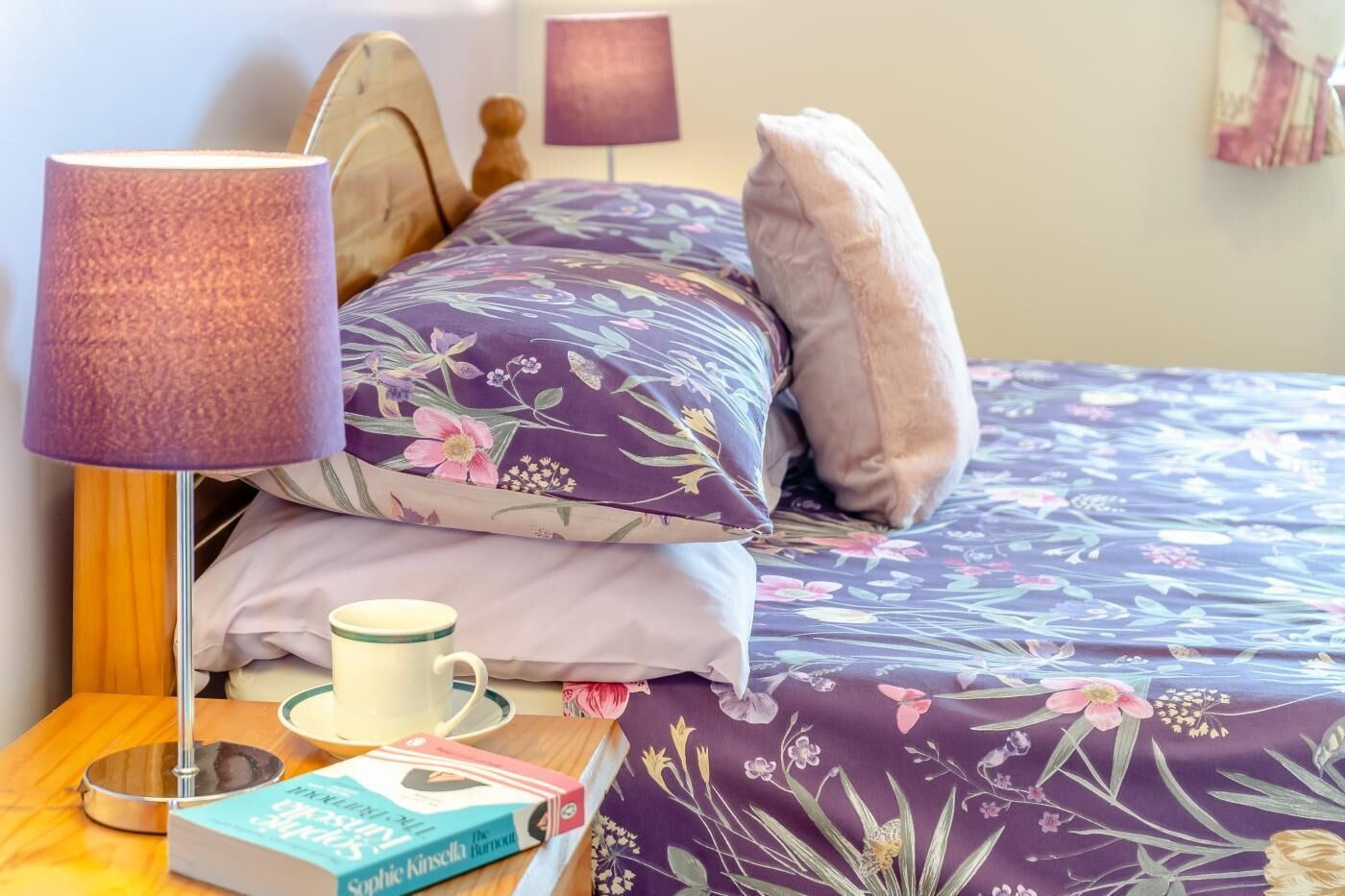 Una cama bien hecha con ropa de cama floral, una lámpara de noche, una taza y un libro sobre una mesita de madera.