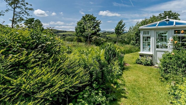 A conservatory with a glass roof, surrounded by lush greenery and a clear blue sky. - 9 Bed in Brecon (oc-bn222) (Sennybridge)