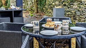 A round glass table with plates, cups, and biscuits, set outdoors with wicker chairs and a stone wall in the background. - 5 Bed in Cartmel (oc-80332) (Cartmel)