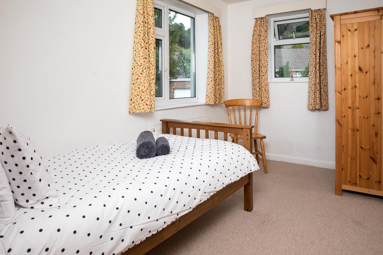 A bedroom with a bed, two pillows, a chair, a wooden wardrobe, and two windows with curtains.