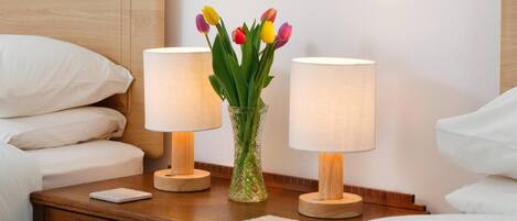 En pænt redt seng med hvide sengetøj, et trænatbord med to bordlamper og en vase med tulipaner.