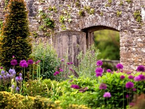 Une arche en pierre surmontée d’une porte en bois, le tout entouré d’une végétation luxuriante et de fleurs violettes.