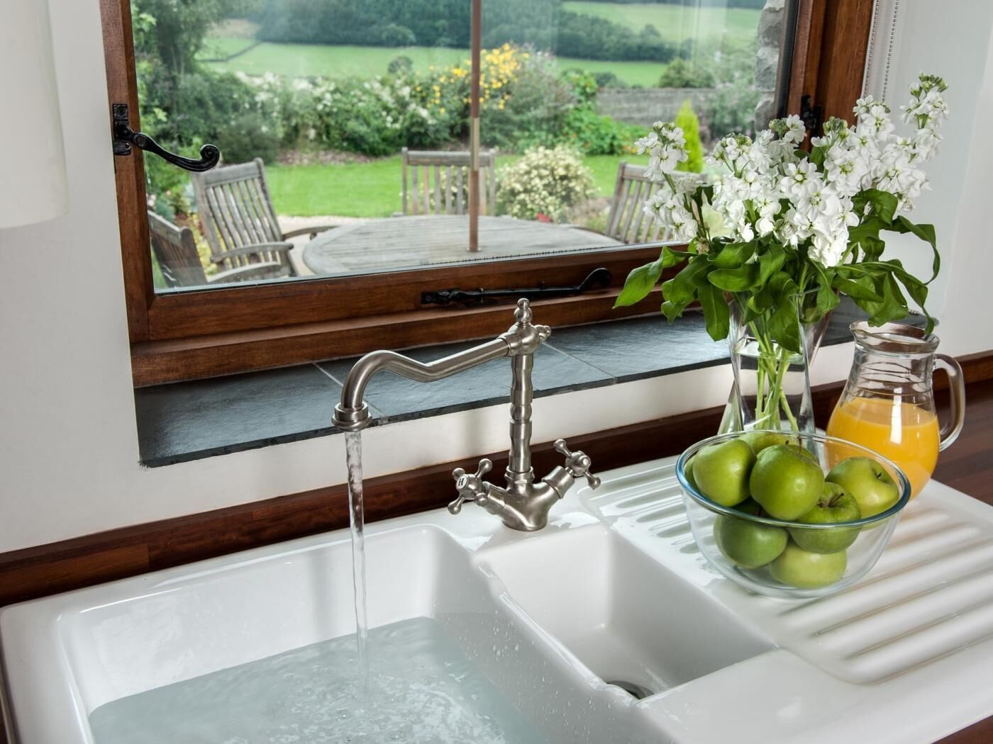 A kitchen sink with running water, a bowl of green apples, and a pitcher of orange juice.
