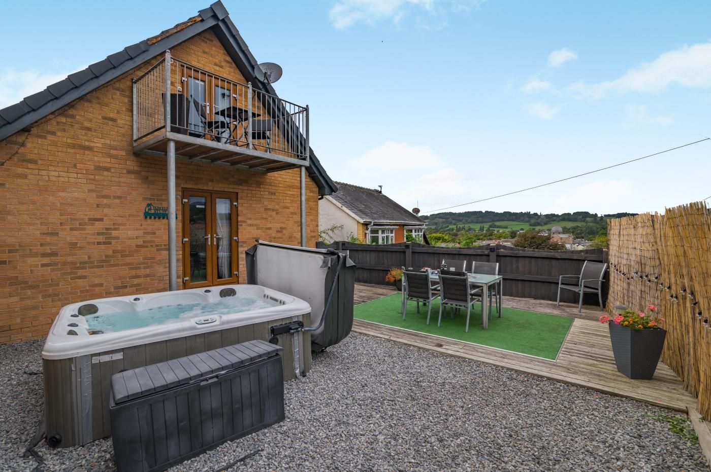 A backyard with a hot tub, a wooden deck, a table with chairs, and a view of the countryside.