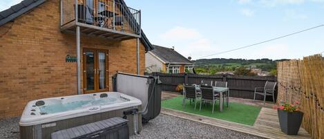 A backyard with a hot tub, a wooden deck, a table with chairs, and a view of the countryside.