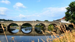 Eine Stebrücke über einem ruhigen Fluss mit klaren Spiegelungen, umgeben von Grün und einem Gebäude mit rotem Dach.