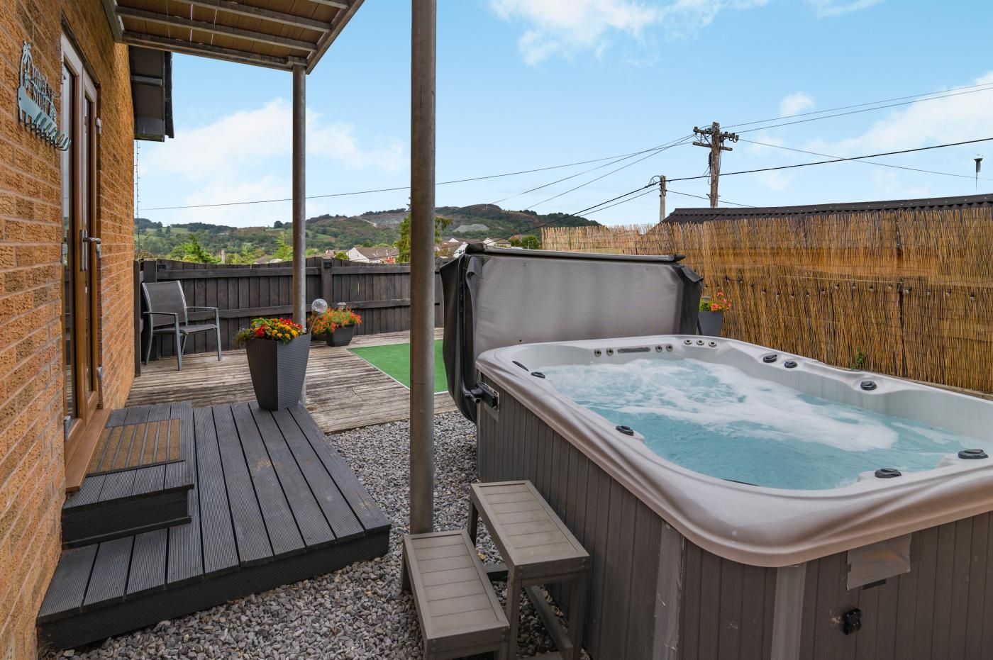 A hot tub in a backyard with a wooden deck, a chair, and potted plants.