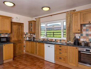 Eine Küche mit Holzausstattung, einem Bosch Geschirrspüler, einem Bosch Backofen, einem Fenster mit Blick auf eine grüne Landschaft und einer Uhr an der Wand.