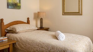 Uma cama bem arrumada com colcha estampada, uma toalha enrolada, uma luminária de cabeceira, uma pequena mesa com livros, um quadro na parede e um espelho.