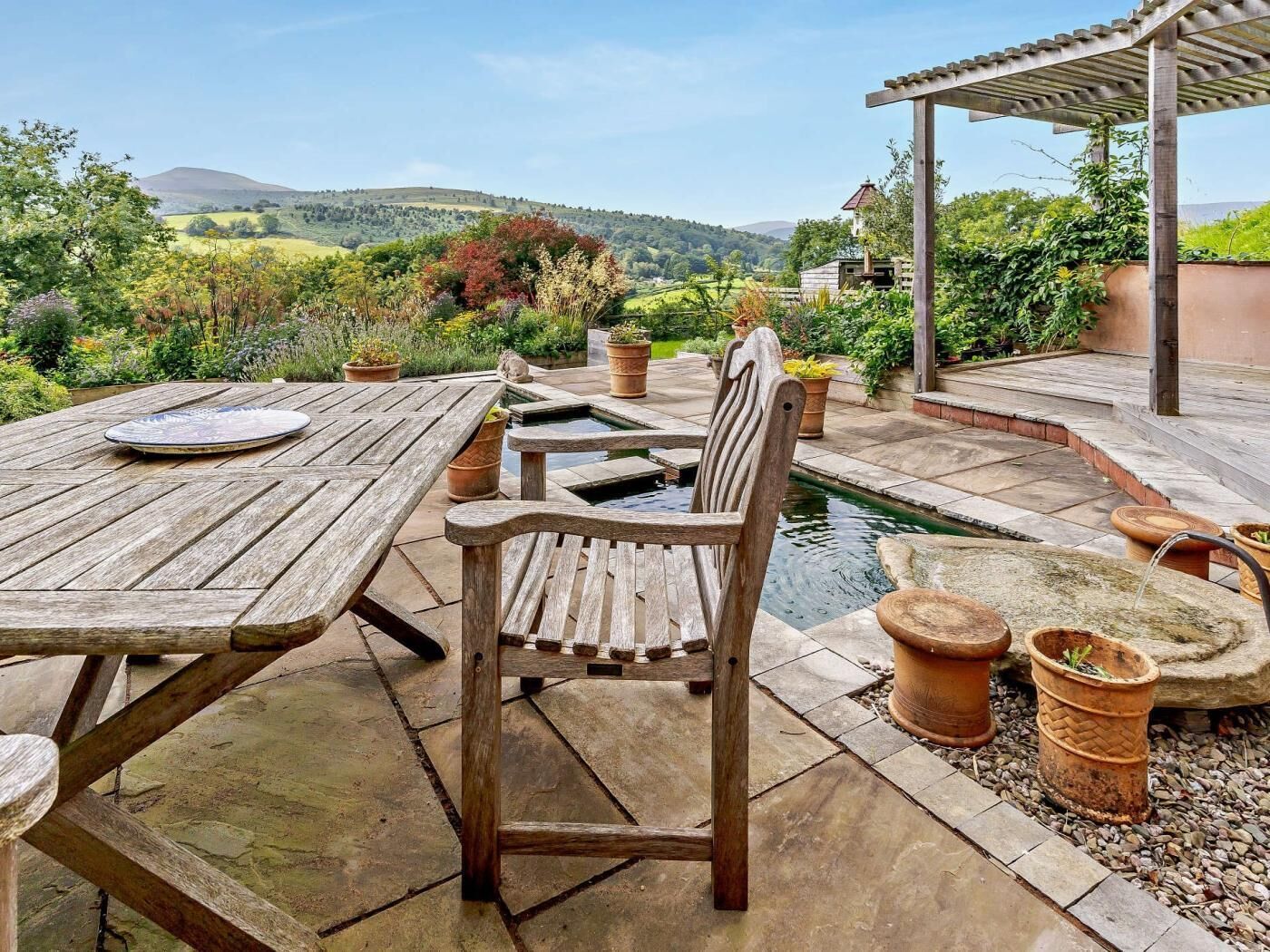 Une terrasse extérieure avec une table et des chaises en bois, entourée de plantes en pot et agrémentée d’un petit élément décoratif aquatique.
