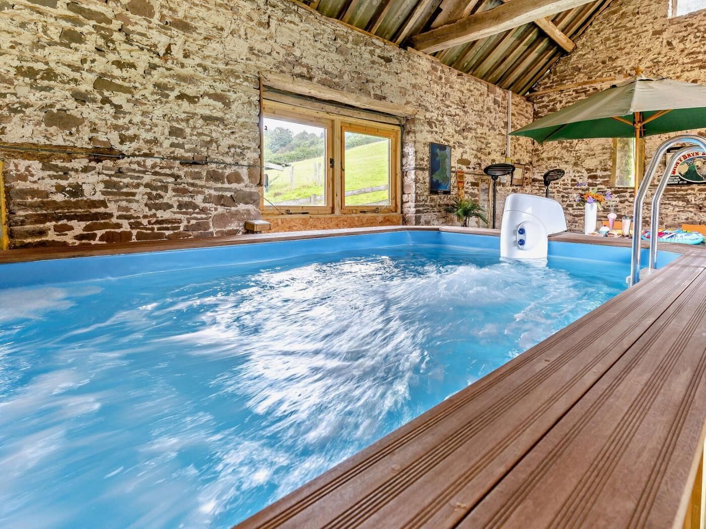Une piscine intérieure aux murs de pierre, dotée d’une terrasse en bois, d’un jacuzzi et d’une fenêtre donnant sur un paysage verdoyant.