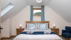 Ein Schlafzimmer mit schrägen Wänden, einem hölzernen Bett, zwei Nachttischen mit Lampen, einem blauen Sessel und einem Fenster mit Vorhängen.