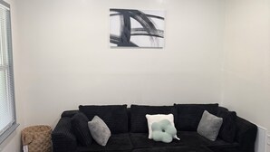 Ein Wohnzimmer mit einem Ecksofa, einem Couchtisch, einem Wandbild und einem Fenster mit Jalousien.