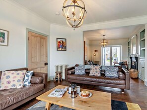 A living room with a brown leather sofa, a wooden coffee table, and a chandelier. - 4 Bed in Tenby (oc-fb193) (Tenby)