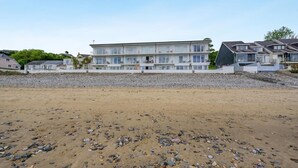 Ein Strandgrundstück mit einem großen Gebäude, das mehrere Balkone aufweist, ein Sandstrand mit vereinzelten Kieselsteinen und ein klarer Himmel.