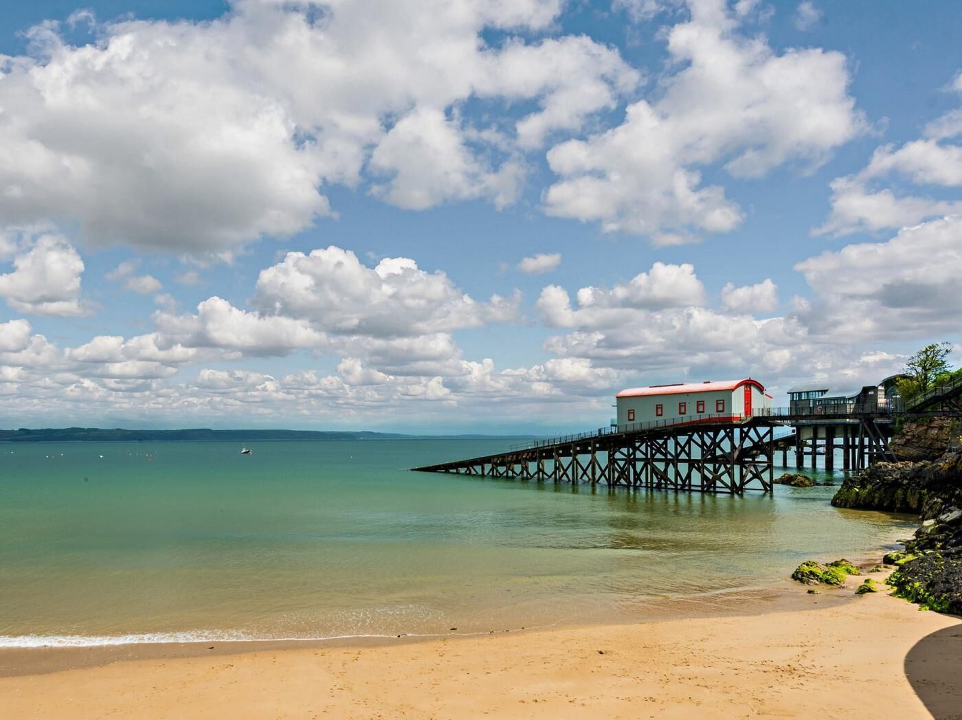A coastal scene with a pier extending into calm waters, a sandy beach, and a building with a red roof on the pier.