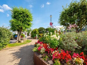 Un sendero en un jardín con diversas flores y árboles, un farol y una persona sentada en un banco.