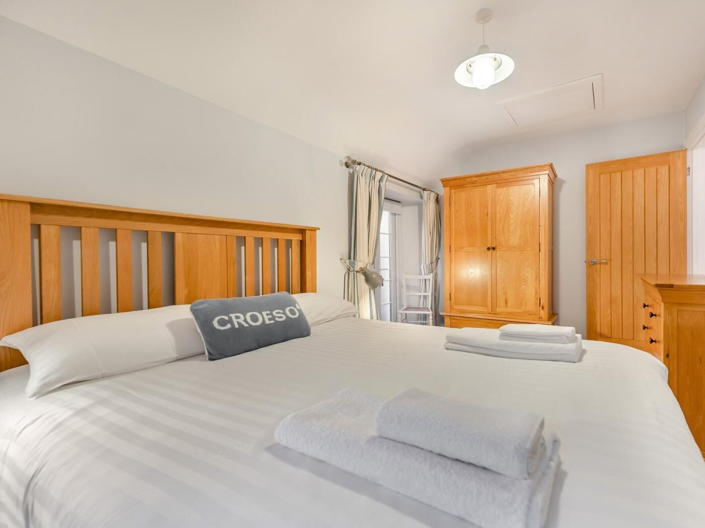 A neatly made bed with a pillow labeled 'CROESO,' a wooden wardrobe, and a chair by the window.