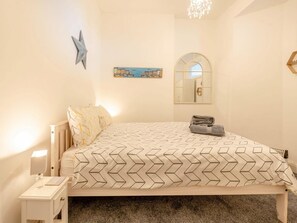 A bedroom with a bed, bedside table, lamp, star decoration, cityscape painting, mirror, and a chandelier. - 2 Bed in Tenby (oc-fb205) (Tenby)